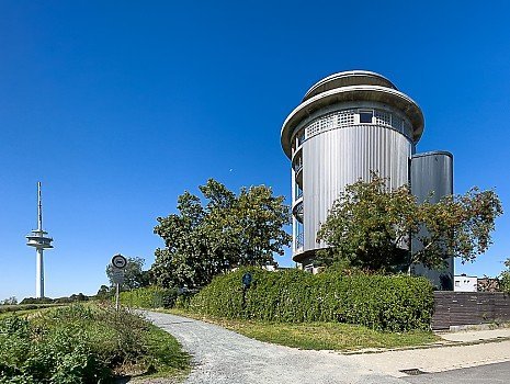 Blick auf den markanten, architektonisch modernisierten Wasserturm in Braunschweig-Broitzem unter strahlend blauem Himmel. Im Hintergrund ragt der wei�-rote Fernsehturm Broitzem auf. Ein Schotterweg f�hrt im Vordergrund an gr�nen Hecken und B�umen vorbei zum Geb�ude.