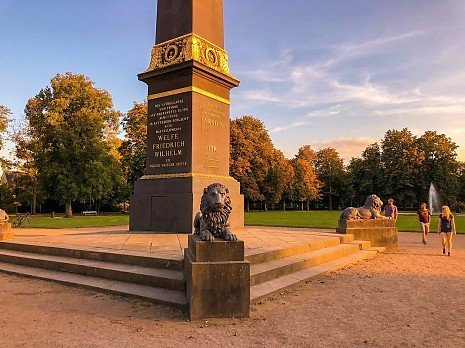 Obelisk mit L�wenstatuen am L�wenwall in Braunschweig bei warmem Abendlicht, umgeben von Parkanlage und Spazierg�ngern im Fr�hling