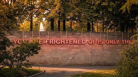 Eine beleuchtete Schriftinstallation an einer Steinmauer im Park L�wenwall in Braunschweig, umgeben von B�umen im warmen Abendlicht; im Hintergrund ist ein Springbrunnen sichtbar.