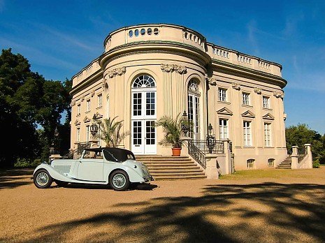 Das Schloss Richmond in Braunschweig an einem sonnigen Tag, ein elegantes klassizistisches Geb�ude mit S�ulen und Rundbau; davor steht ein hellblauer Oldtimer auf einer Kiesfl�che, umgeben von park�hnlicher Anlage.