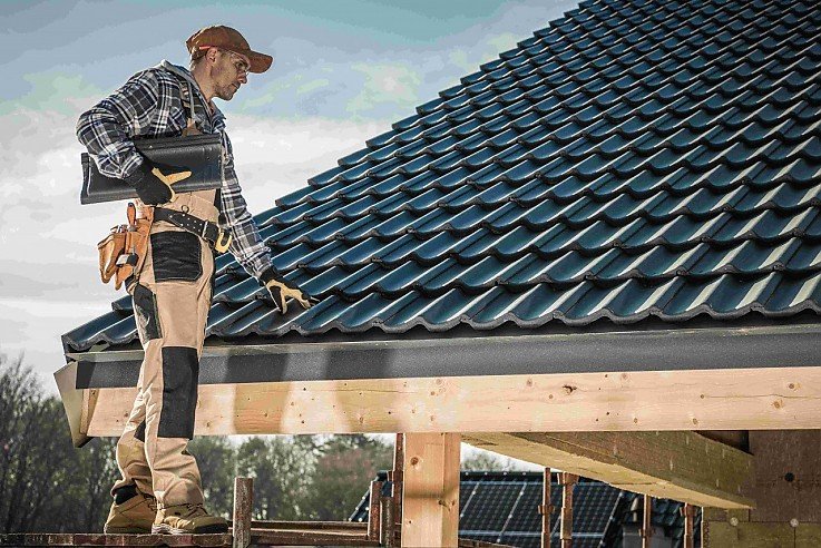 Ein Handwerker in Arbeitskleidung steht auf einem Gerüst an einem neu gedeckten, dunklen Hausdach und prüft die Dachziegel im Zuge einer Dachsanierung. Ein Handwerker in Arbeitskleidung steht auf einem Gerüst an einem neu gedeckten, dunklen Hausdach und prüft die Dachziegel im Zuge einer Dachsanierung.